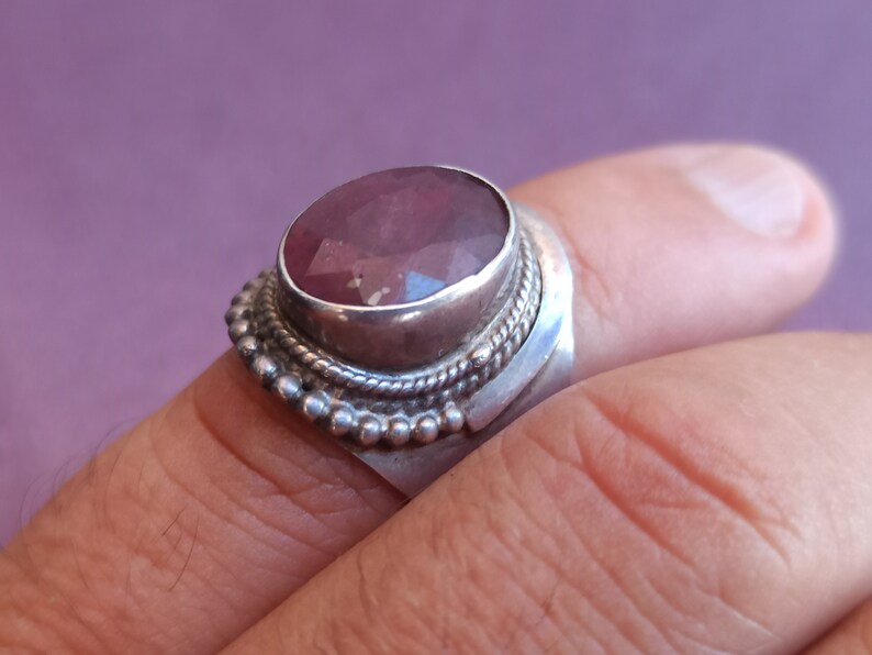 Pode incluir: Um anel de prata com uma grande gema oval rosa engastada em um engaste de bezel. O anel tem uma banda decorativa com uma borda de contas.