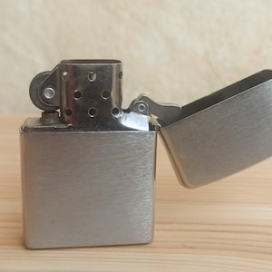K&ouml;nnte beinhalten: Ein silberner Feuerzeug mit geb&uuml;rsteter Metalloberfl&auml;che. Das Feuerzeug ist ge&ouml;ffnet und der Deckel ist nach hinten geklappt.