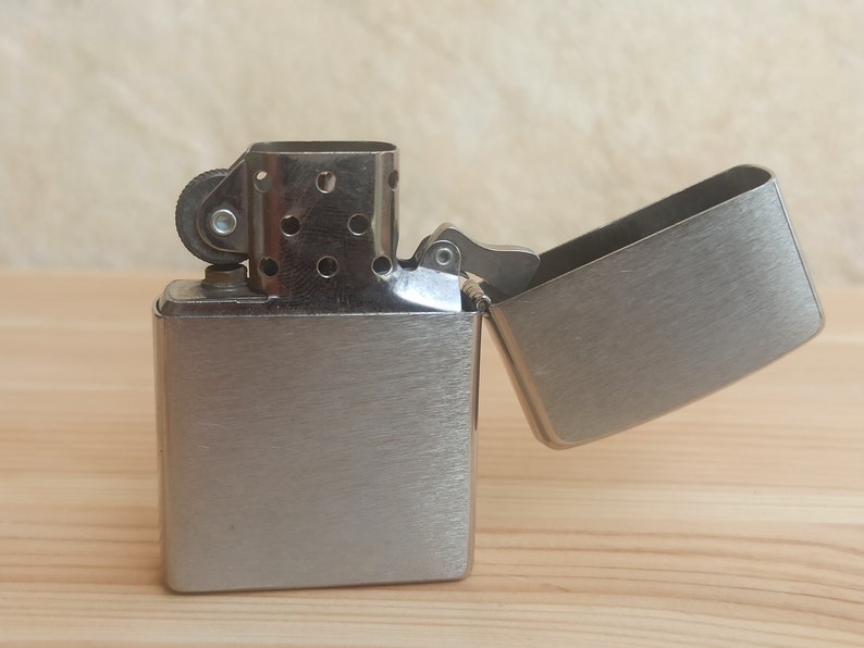 K&ouml;nnte beinhalten: Ein silberner Feuerzeug mit geb&uuml;rsteter Metalloberfl&auml;che. Das Feuerzeug ist ge&ouml;ffnet und der Deckel ist nach hinten geklappt.