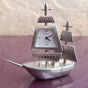 May include: A silver-colored ship-shaped clock with a white clock face. The clock has a quartz movement and the clock face displays the time. The clock has two masts and a long, thin bowsprit.