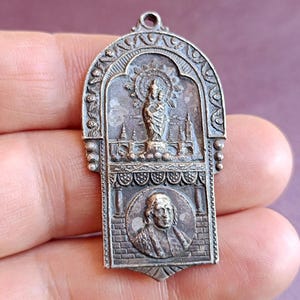 Peut inclure: Pendentif argenté avec une partie supérieure arquée, représentant une figure religieuse et un portrait. Le pendentif présente des détails complexes, notamment des éléments floraux et architecturaux. Le pendentif est tenu dans une main.