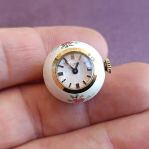 May include: A small, round vintage watch with a cream-colored face and gold-tone bezel. The watch face has black hands and hour markers. The watch is decorated with small pink and green floral designs. The watch measures approximately 1 inch in diameter.