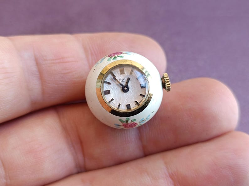 May include: A small, round vintage watch with a cream-colored face and gold-tone bezel. The watch face has black hands and hour markers. The watch is decorated with small pink and green floral designs. The watch measures approximately 1 inch in diameter.
