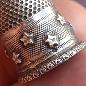 May include: Close-up of a silver thimble with a textured surface and star-shaped embellishments. The thimble has a perforated top and decorative bands. The design is detailed and the metal has a slightly aged appearance.