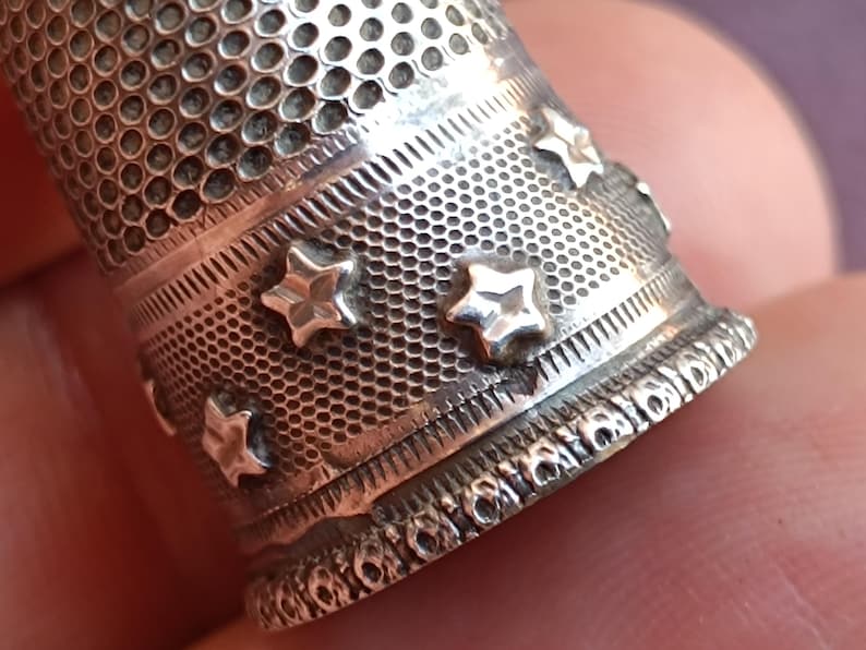 May include: Close-up of a silver thimble with a textured surface and star-shaped embellishments. The thimble has a perforated top and decorative bands. The design is detailed and the metal has a slightly aged appearance.