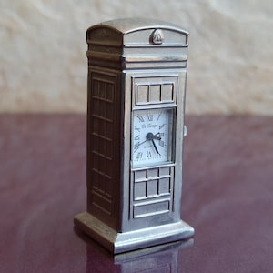 Può includere: Una replica in miniatura color argento di una cabina telefonica britannica con un quadrante dell'orologio che recita "El Tiempo" e "Quartz".