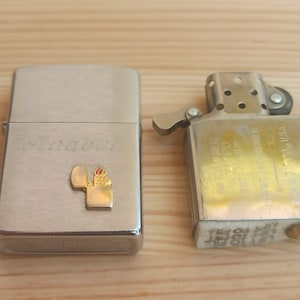 K&ouml;nnte beinhalten: Zwei silberne Feuerzeuge, eines mit einer personalisierten Inschrift "Anabel" und einem kleinen goldenen Flammenlogo, das andere mit einer goldenen Oberfl&auml;che und verschiedenen Texten in Japanisch und Englisch.
