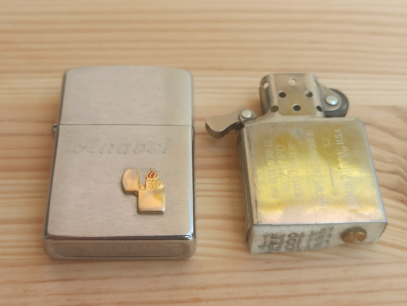 K&ouml;nnte beinhalten: Zwei silberne Feuerzeuge, eines mit einer personalisierten Inschrift "Anabel" und einem kleinen goldenen Flammenlogo, das andere mit einer goldenen Oberfl&auml;che und verschiedenen Texten in Japanisch und Englisch.
