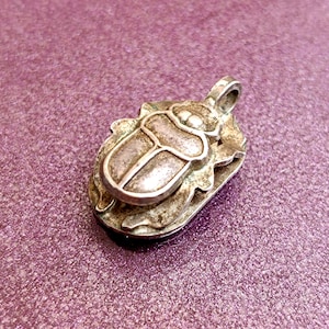 May include: A silver-toned pendant in the shape of a scarab beetle, featuring detailed etching. The pendant has a loop for a chain and is set against a purple background. The beetle design is raised and has a textured appearance.