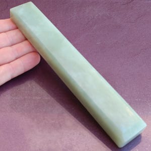 May include: A pale green, rectangular sharpening stone with beveled edges. The stone is held by a hand, and the background is a muted purple color. The stone appears to be made of jade or a similar material.