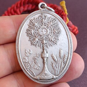 May include: An oval silver-toned metal pendant featuring a raised relief of a monstrance at the center. The design includes grapevines with clusters of grapes on the left and stalks of wheat on the right. A small metal loop is attached to the top.