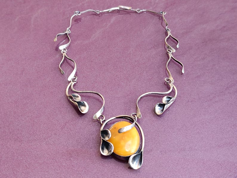 May include: A silver necklace with a large amber pendant. The pendant is surrounded by silver leaves and the necklace is made of a series of silver loops.