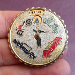 May include: A vintage round badge with a gold-coloured scalloped edge. The badge features a central traffic officer surrounded by a red motorcycle, blue car, red car, and green scooter. The word "BADEN" is at the top.