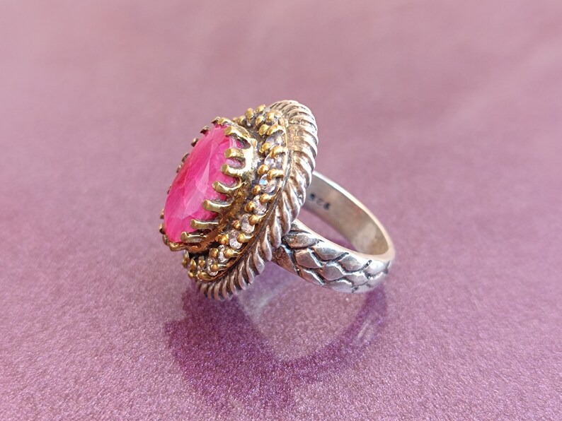Op de afbeelding: Een zilveren ring met een roze edelsteen gezet in een gouden setting. De ring heeft een gevlochten band.