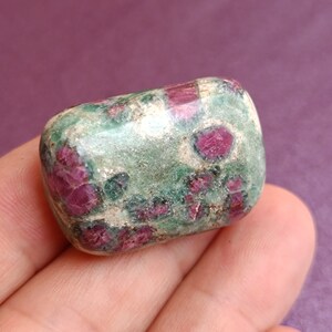 May include: A rounded, polished gemstone with a mix of colours. The stone has a green base with patches of deep red. The shape is smooth and tumbled, giving it a polished appearance. Likely a ruby gemstone.