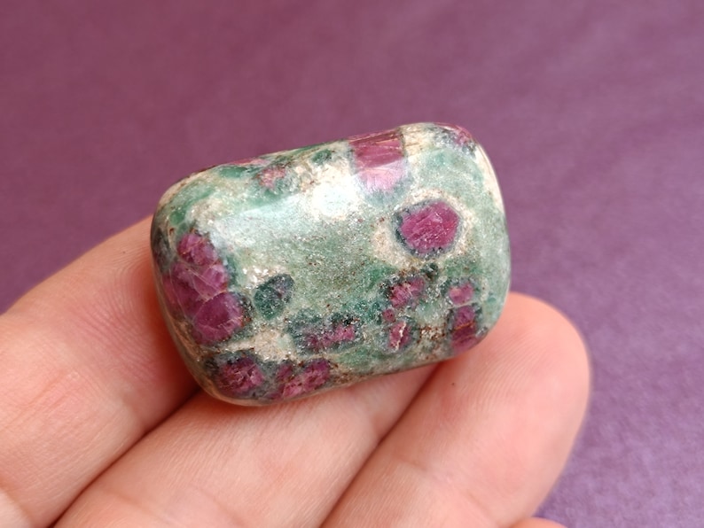 May include: A rounded, polished gemstone with a mix of colours. The stone has a green base with patches of deep red. The shape is smooth and tumbled, giving it a polished appearance. Likely a ruby gemstone.