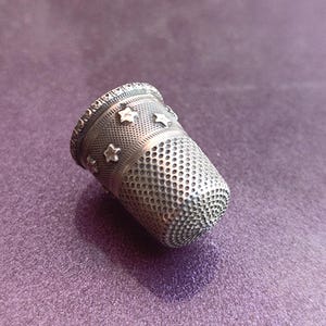 May include: A silver thimble with a decorative star pattern. The thimble has a textured surface with small holes and a detailed rim. The sewing tool is set against a purple background.