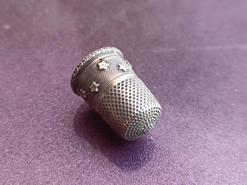 May include: A silver thimble with a decorative star pattern. The thimble has a textured surface with small holes and a detailed rim. The sewing tool is set against a purple background.