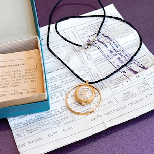 Puede incluir: Un reloj de bolsillo dorado con un collar de cordón negro. El reloj está en una caja redonda con una lupa alrededor de la esfera. El reloj está sobre un papel blanco con texto negro. El texto incluye las palabras "Код ОКП" y "150988".