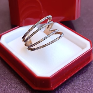 May include: A gold-toned ring with three curved bands, each encrusted with small, iridescent stones. The ring is displayed in a red and white jewelry box, creating a striking contrast. The ring's design is modern and elegant.