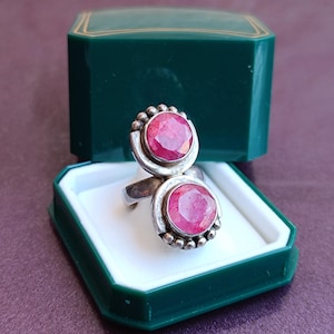 Puede incluir: Un anillo de plata con dos grandes piedras preciosas rosas brillantes de corte ovalado. El anillo se muestra en un joyero verde oscuro con interior blanco. Las piedras est&aacute;n engastadas en plata con peque&ntilde;os detalles redondos.