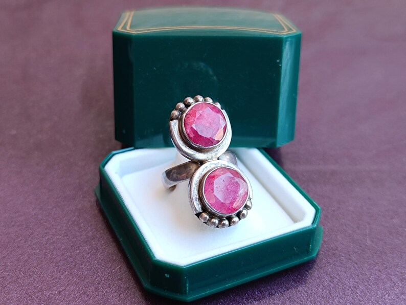 Puede incluir: Un anillo de plata con dos grandes piedras preciosas rosas brillantes de corte ovalado. El anillo se muestra en un joyero verde oscuro con interior blanco. Las piedras est&aacute;n engastadas en plata con peque&ntilde;os detalles redondos.