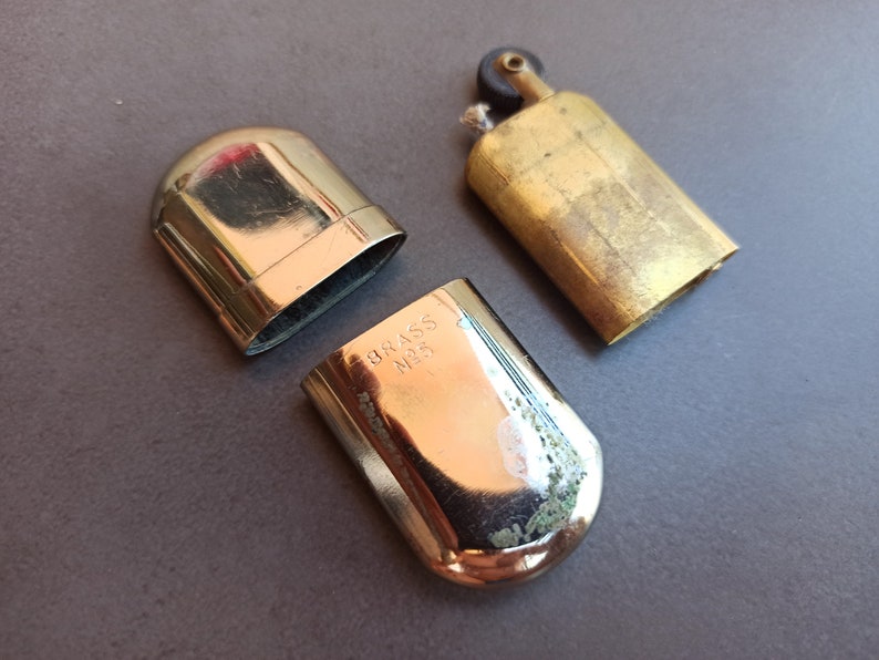 May include: A vintage brass lighter with a hinged lid and a small, round knob on the top. The lighter is engraved with the words "BRASS NO. 5".