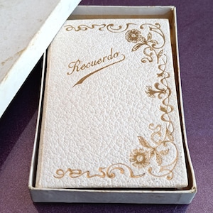 May include: A white leather-bound journal with gold floral designs and the word "Recuerdo" written in cursive on the front cover.