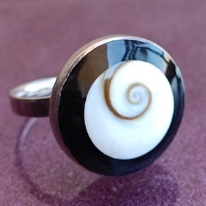 May include: A silver ring featuring a large, circular design. The ring's centerpiece is a white spiral set against a black background, encased in a silver band. The ring is displayed on a purple surface.