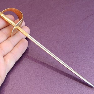 K&ouml;nnte beinhalten: Ein goldfarbenes Metalls&auml;bel mit einer silbernen Klinge. Das S&auml;bel ist etwa 25 cm lang und hat einen verzierten Griff.
