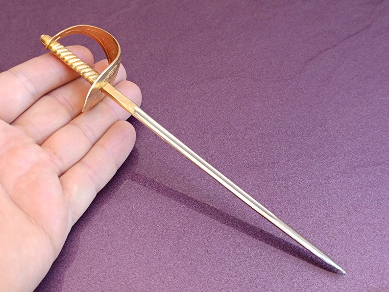K&ouml;nnte beinhalten: Ein goldfarbenes Metalls&auml;bel mit einer silbernen Klinge. Das S&auml;bel ist etwa 25 cm lang und hat einen verzierten Griff.
