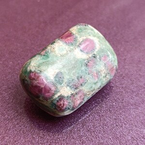 May include: A rounded gemstone with a mix of colours. The stone has a green base, speckled with deep red patches and some white. The stone is smooth and polished. The background is a solid, textured purple.