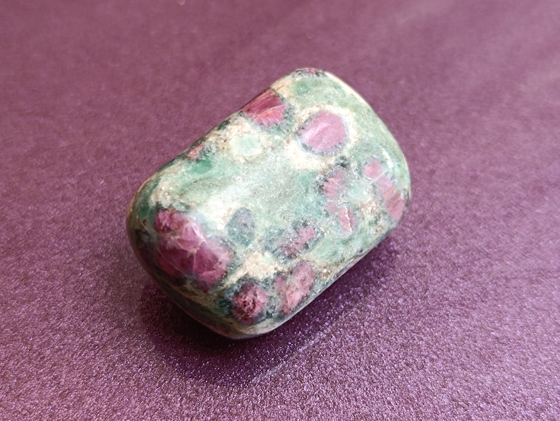 May include: A rounded gemstone with a mix of colours. The stone has a green base, speckled with deep red patches and some white. The stone is smooth and polished. The background is a solid, textured purple.