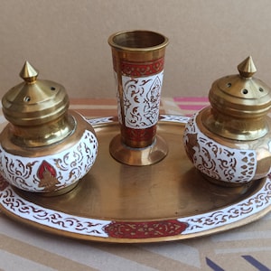 May include: A set of three gold-toned metal containers with white and red painted designs. The containers are on a matching tray with a similar design. The containers have a small opening on the top and may be used for spices or condiments.