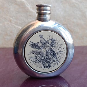 Peut inclure: Une gourde en métal argenté de forme ronde et plate. La gourde a un bouchon à vis et un panneau circulaire blanc avec une illustration en noir et blanc d'un faisan.