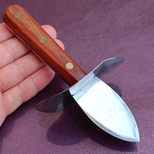 May include: An oyster knife with a wooden handle and a stainless steel blade. The handle is a dark brown color with three brass rivets. The blade is curved and has the text "RUFF" etched on it.