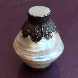 Può includere: Un piccolo vaso decorativo bianco, iridescente, madreperlaceo, con un coperchio argentato e una fascia argentata con motivi floreali attorno alla parte superiore.