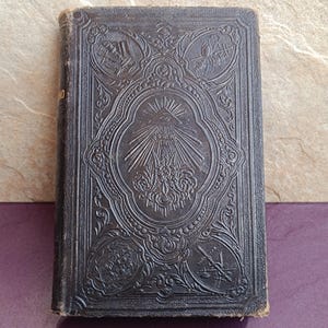 Könnte beinhalten: Antikes, dunkelbraunes, ledergebundenes Buch mit aufwendigen geprägten Mustern. Das Cover zeigt ein zentrales ovales Design mit einem Auge und strahlenförmigen Linien, umgeben von anderen dekorativen Elementen. Die Kanten des Buches weisen Gebrauchsspuren auf.