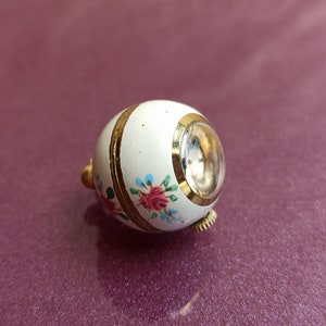May include: A small, white, egg-shaped trinket box with a clear, circular window. The box is decorated with gold bands and floral designs in pink, blue, and green. The trinket box has a gold-colored clasp.