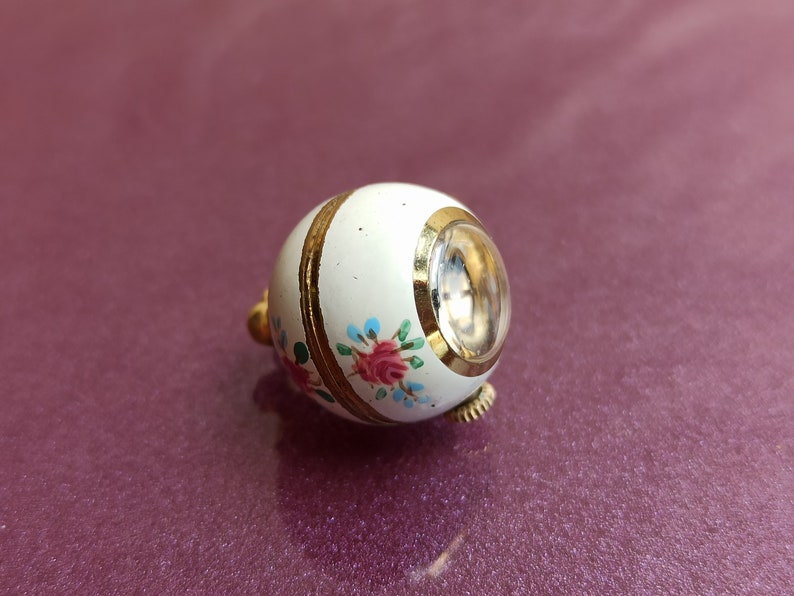 May include: A small, white, egg-shaped trinket box with a clear, circular window. The box is decorated with gold bands and floral designs in pink, blue, and green. The trinket box has a gold-colored clasp.