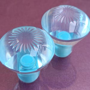 Op de afbeelding: Twee heldere, lichtblauwe glazen kastknoppen. Elke knop heeft een rond, geëtst bloemmotief en een bijpassende lichtblauwe basis. De knoppen staan tegen een paarse achtergrond.