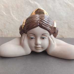 May include: A ceramic figurine of a young girl with brown hair and blue eyes. She is lying on her stomach with her hands under her chin. Her hair is adorned with gold accents.