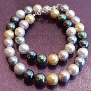 May include: A multi-strand pearl necklace featuring a variety of colors, including white, silver, gold, and dark green. The pearls are round and strung together with a silver clasp. The necklace is displayed on a purple surface.