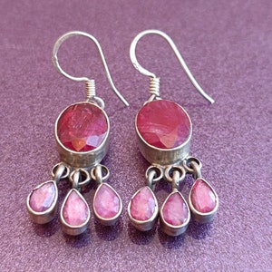 May include: A pair of silver dangle earrings with pink gemstone drops. The earrings feature a large oval-shaped pink gemstone at the top and three smaller teardrop-shaped pink gemstones dangling below.