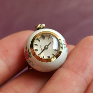 May include: A small, vintage, round watch with a white face and gold-tone bezel. The watch has a floral design in pink, red, and blue. The watch face reads "lana". The watch is held in a hand.
