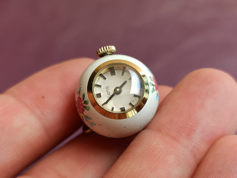 May include: A small, vintage, round watch with a white face and gold-tone bezel. The watch has a floral design in pink, red, and blue. The watch face reads "lana". The watch is held in a hand.