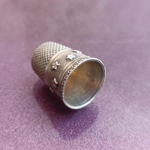 May include: A silver thimble with a textured surface and star-shaped embellishments. The thimble is designed for sewing and crafting, with a detailed pattern around the top and a smooth interior.