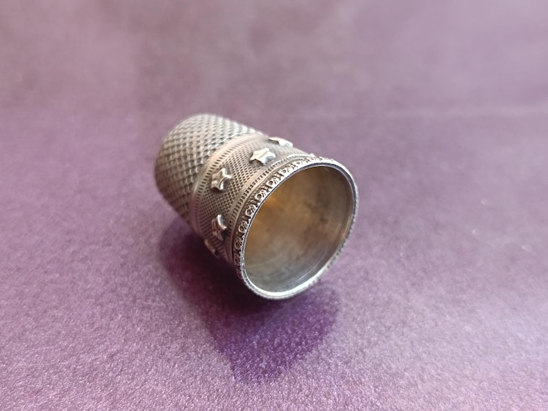 May include: A silver thimble with a textured surface and star-shaped embellishments. The thimble is designed for sewing and crafting, with a detailed pattern around the top and a smooth interior.