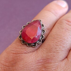 Pode incluir: Um anel de prata com uma grande pedra vermelha em forma de l&aacute;grima engastada em uma moldura de prata decorativa.