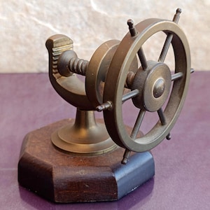 May include: A brass ship wheel mounted on a threaded metal spindle and a curved support arm, attached to a dark brown octagonal wooden base. The metallic components feature a muted, aged finish with visible screw threads on the central rod.